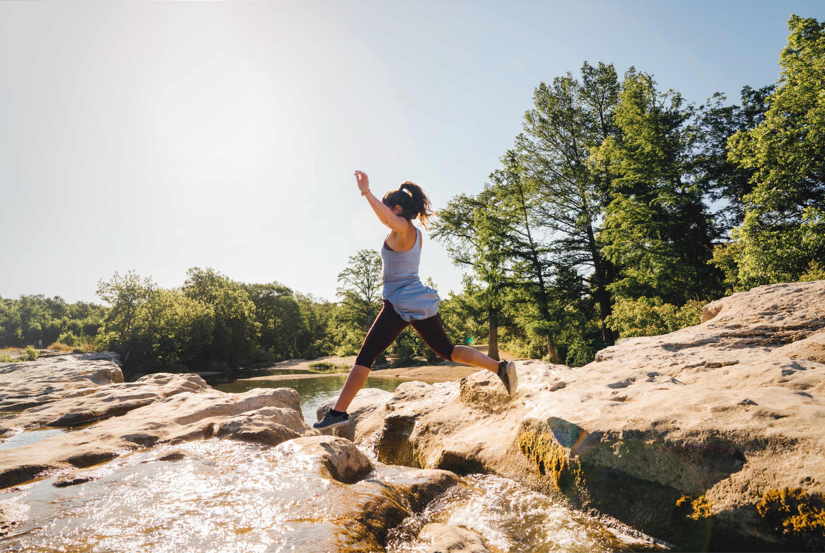 A trail runner hops joyfully along boulders surrounding a woodland stream. The sun-kissed water glistens, and the treetops glow vibrantly under a clear blue sky.